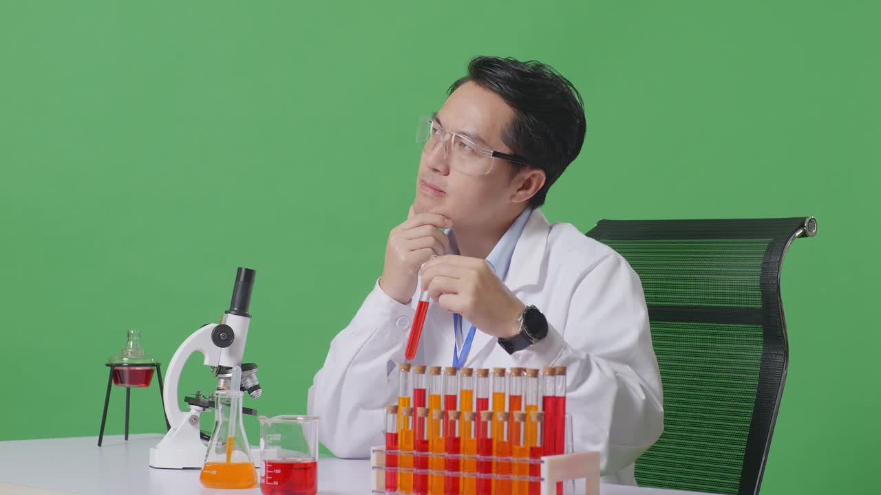 아시아 남성 과학자가 실험관에서 생각하고, 주위를 둘러보고, 지표손가락을 올리며 녹색 스크린 백그라운드 실험실에서 현미경으로 테이블에서 작업합니다.