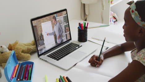 african american girl doing homework while having a video call with male teacher on laptop at home