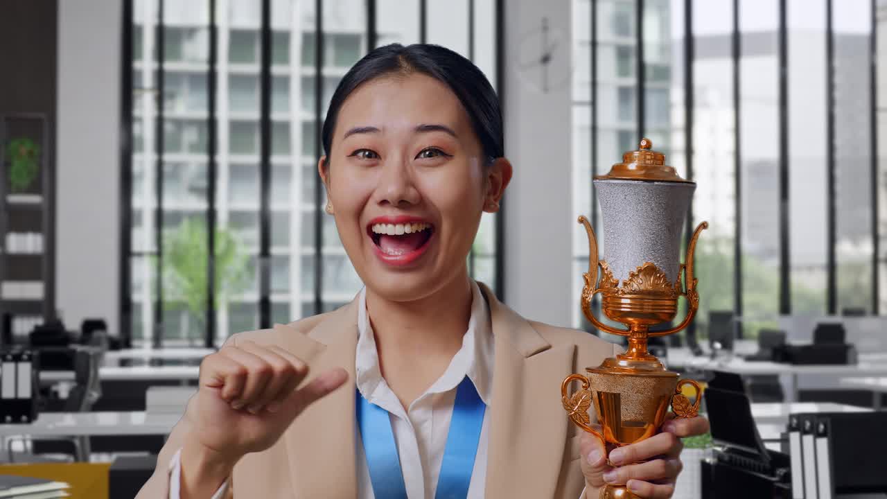 Close Up Of Asian Business Woman In A Suit With A Gold Medal Showing Gold Trophy To Camera, Smiling, And Pointing Herself Being Proud Winning As The First Winner In The Office