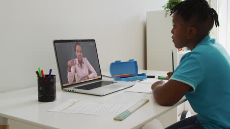 african american boy doing homework while having a video call with female teacher on laptop at home