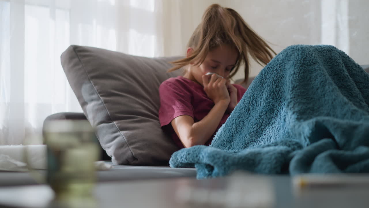 Primo piano di una tazza di vetro con bambino sdraiato sul divano con l'influenza, a riposo sotto una coperta con un fazzoletto in mano, che mostra segni di malattia, debolezza e disagio dovuti ai sintomi del raffreddore o dell'influenza