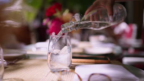 pouring sparkling water from a glass bottle into a drinking glass with a table setting in the background
