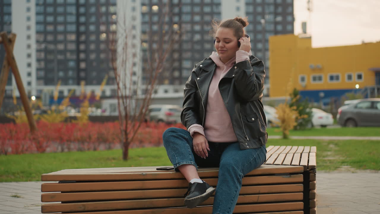 Music artist seated on wooden bench in urban park, while holding headset and nodding rhythmically to music surrounded by greenery parked cars and tall buildings in background