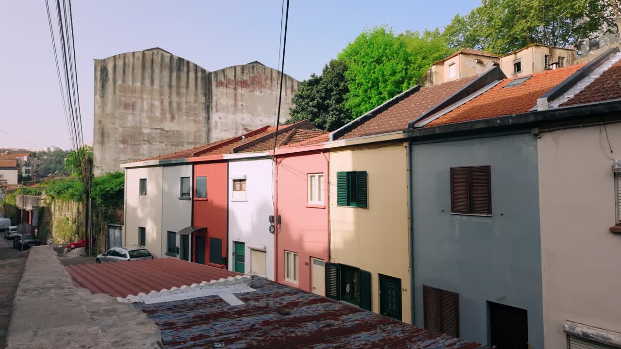Pintoresca hilera de casas tradicionales de Oporto pintadas en varios colores, adornadas con tejados de tejas clásicas. Líneas eléctricas aéreas y un gran edificio industrial forman el telón de fondo.