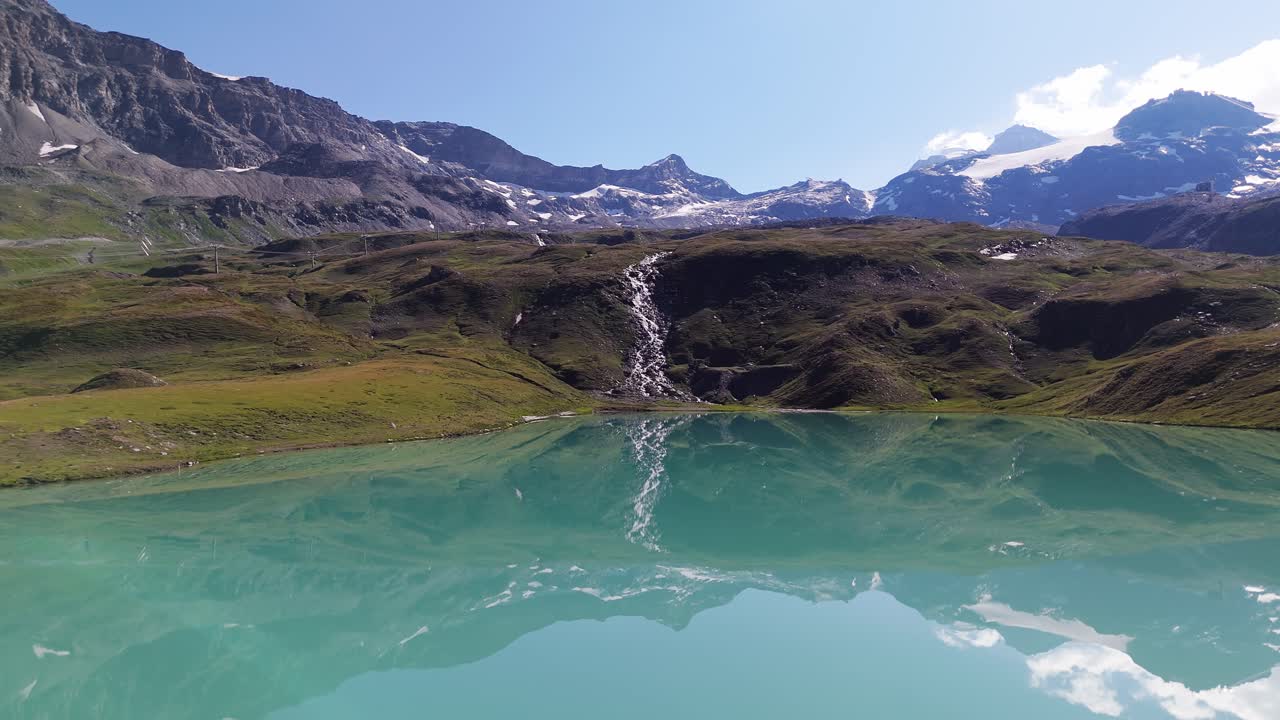 Aerial drone shot of a beautiful blue-green lake in Cervinia, Italy, with a waterfall and summer vibes