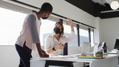 two diverse female colleagues wearing face masks looking at laptop and discussing in office