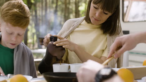 smiling woman pouring coffee into a mug while having breakfast with her family at camping in the forest