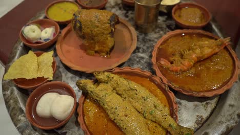 cinematic shot of multiple dishes like fishes, rasgullas , papad, sandesh , prawns , kheer and some other dishes given on clay plates and kept on large aluminum plate