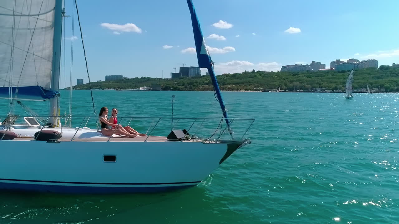 People rest on a luxury yacht. Sailboat under white sail floating on the open sea. Beautiful yacht sail on a seascape. Women relax on boat at summer cruise. Camera rising.