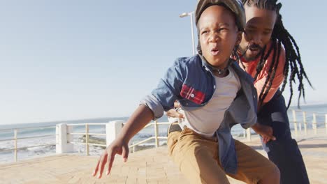 video de un feliz padre afroamericano aprendiendo a su hijo a patinar en el paseo marítimo
