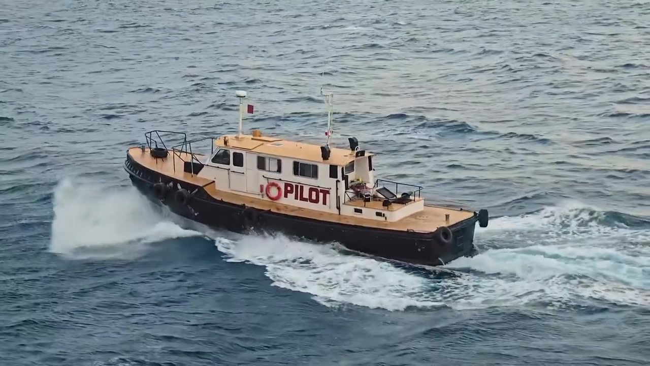 A specialized pilot boat with a dark hull and beige cabin navigates quickly through choppy blue ocean waters, creating a white wake and splashing waves.