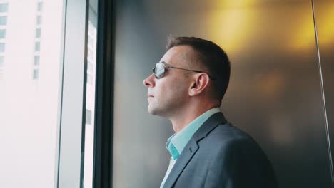 Modern-businessman-rising-in-lift-and-looking-to-skyscrapers.-Man-in-elevator