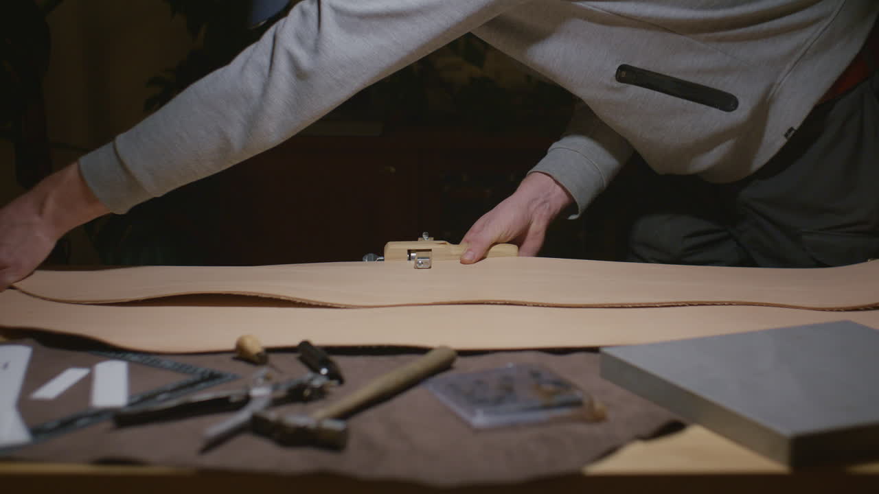 A craftsman in a gray sweatshirt cuts a piece of leather on a workbench, surrounded by various leatherworking tools in a dimly lit workshop.