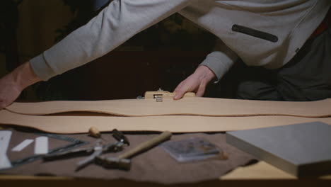 a craftsman in a gray sweatshirt cuts a piece of leather on a workbench, surrounded by various leatherworking tools in a dimly lit workshop.
