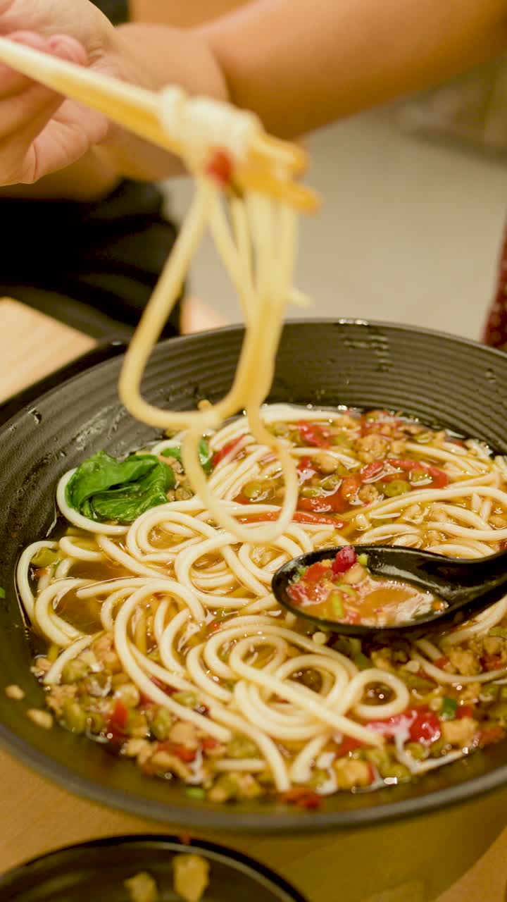 Una persona solleva zuppa di noodle piccante con le bacchette in un ambiente da pranzo illuminato caldamente, in prospettiva ravvicinata.