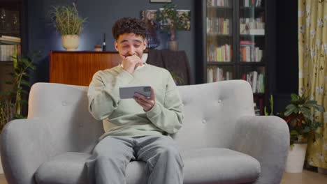 young man watching some unexpected movie scene on smartphone while sitting on couch at home