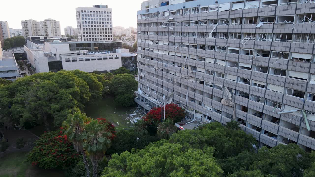 Drone pullback, damaged residential building, broken windows, northern Tel Aviv, missile aftermath, urban area, building exterior, green trees, impact damage, visible debris