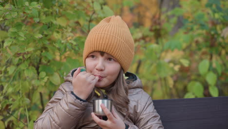 kid dressed in warm coat and orange knit hat gently blows on warm drink to cool it, then dips finger to test temperature while sitting on bench surrounded by colorful autumn foliage