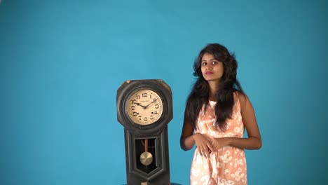 Ein-Junges-Indisches-Mädchen-In-Orangefarbenem-Kittel,-Das-Mit-Einer-Alten-Vintage-Uhr-Auf-Einem-Isolierten-Blauen-Hintergrund-Steht