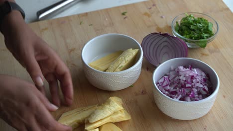 Bonita-Toma-Panorámica-Desde-Arriba-De-Un-Plátano-Amarillo-Maduro-Que-Se-Agrega-A-Un-Tazón-Blanco-E-Ingredientes-Especiales-Para-Cocinar-Una-Comida,-Dos-Latas-De-Frijoles,-Arroz,-Plátano,-Aguacate,-Cebolla-Roja-Y-Cilantro.