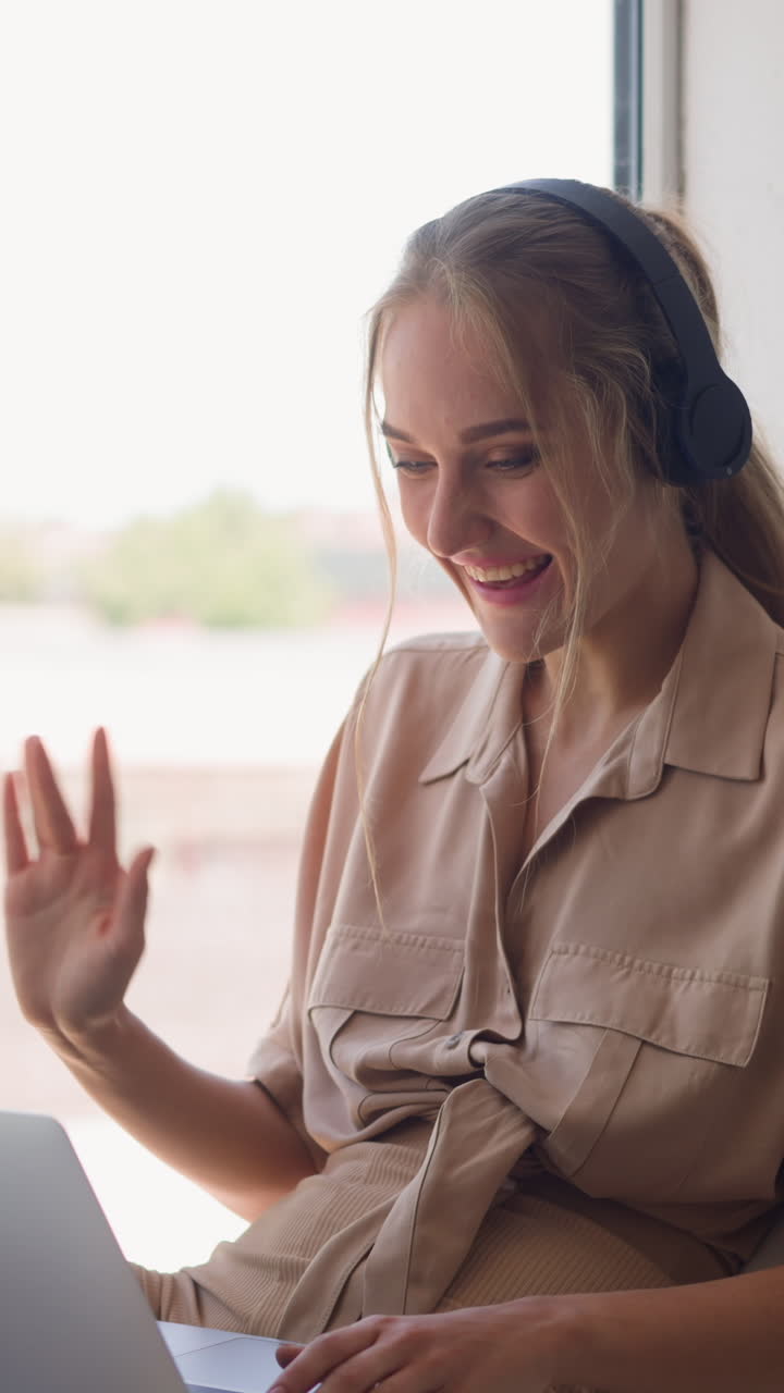 Young woman in headphones waves hand to greet talking on videocall via laptop by window leaned on pillow in comfortable room at home slow motion