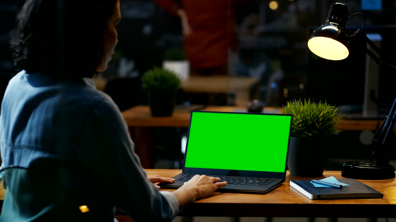 una trabajadora de oficina en su escritorio trabaja en una computadora portátil con una maqueta de pantalla verde. sobre las imágenes del hombro. se sienta en el escritorio de madera en la oficina creativa.