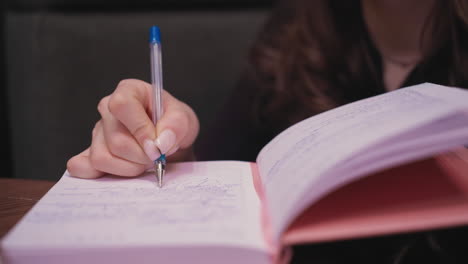 close up of female hand holding blue pen writing in pink notebook with visible ink, soft lighting highlights fingertips and handwriting as she focuses in relaxed indoor setting, showing concentration