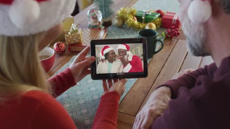 Kaukasisches-Paar-Mit-Weihnachtsmützen-Nutzt-Tablet-Für-Weihnachtsvideoanruf-Mit-Paar-Auf-Dem-Bildschirm