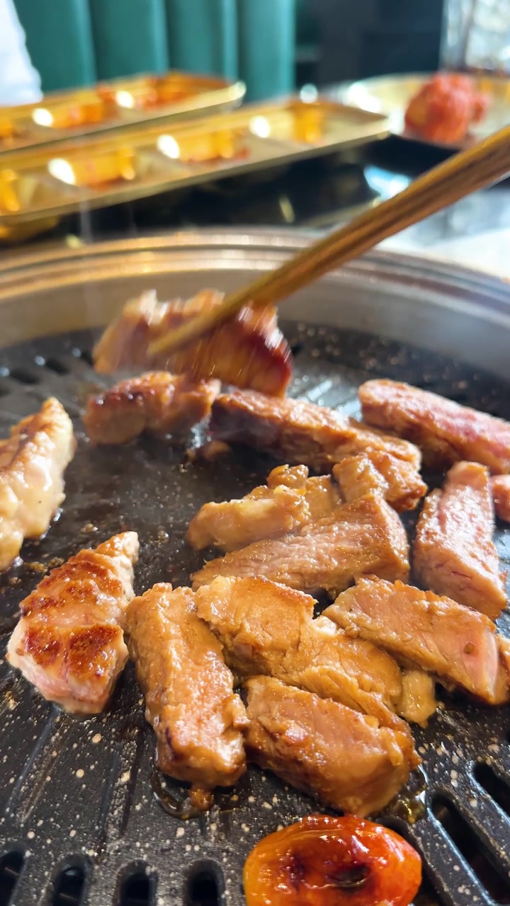 Des baguettes soulèvent des tranches de porc grillé d'un grill de table grésillant dans un restaurant moderne et lumineux.