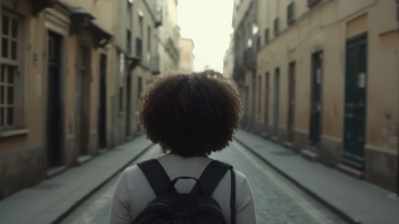 Walking woman wearing light longsleeve top, black backpack moving down cobbled lane to lit horizon