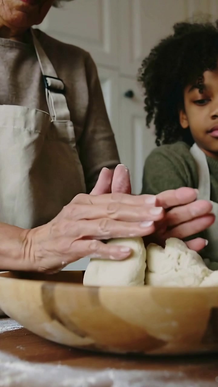 Vertical video: Placing hands on bowl, senior woman showing child kneading dough in kitchen, aprons