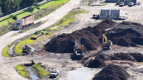 drone captures excavators moving soil in an industrial setting with trucks and greenery under bright daylight