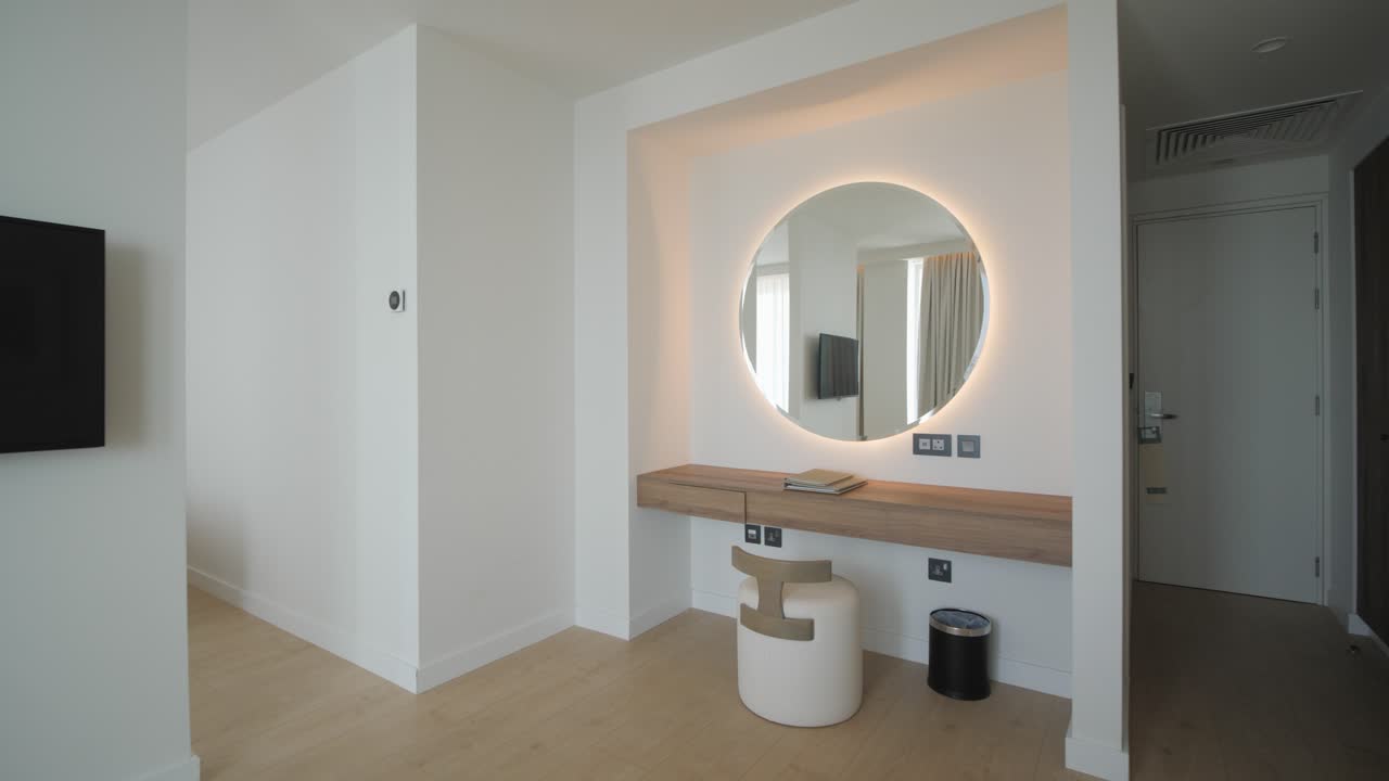 A gimbal shot of an empty luxurious hotel apartment room with TV, sofa, chairs and large round backlit mirror on the wall with sea view
