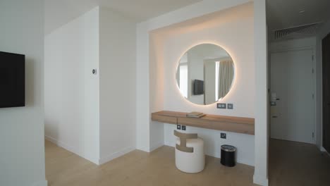 a gimbal shot of an empty luxurious hotel apartment room with tv, sofa, chairs and large round backlit mirror on the wall with sea view