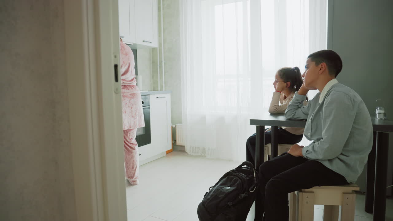 nanny wearing pink pajamas preparing breakfast on kitchen countertop with kettle while two school uniform kids sit at table waiting with backpacks near curtain bright window morning routine scene