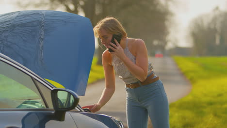 Frustrierte-Frau-Mit-Kaputtem-Auto-Auf-Der-Landstraße,-Die-Per-Mobiltelefon-Um-Hilfe-Ruft