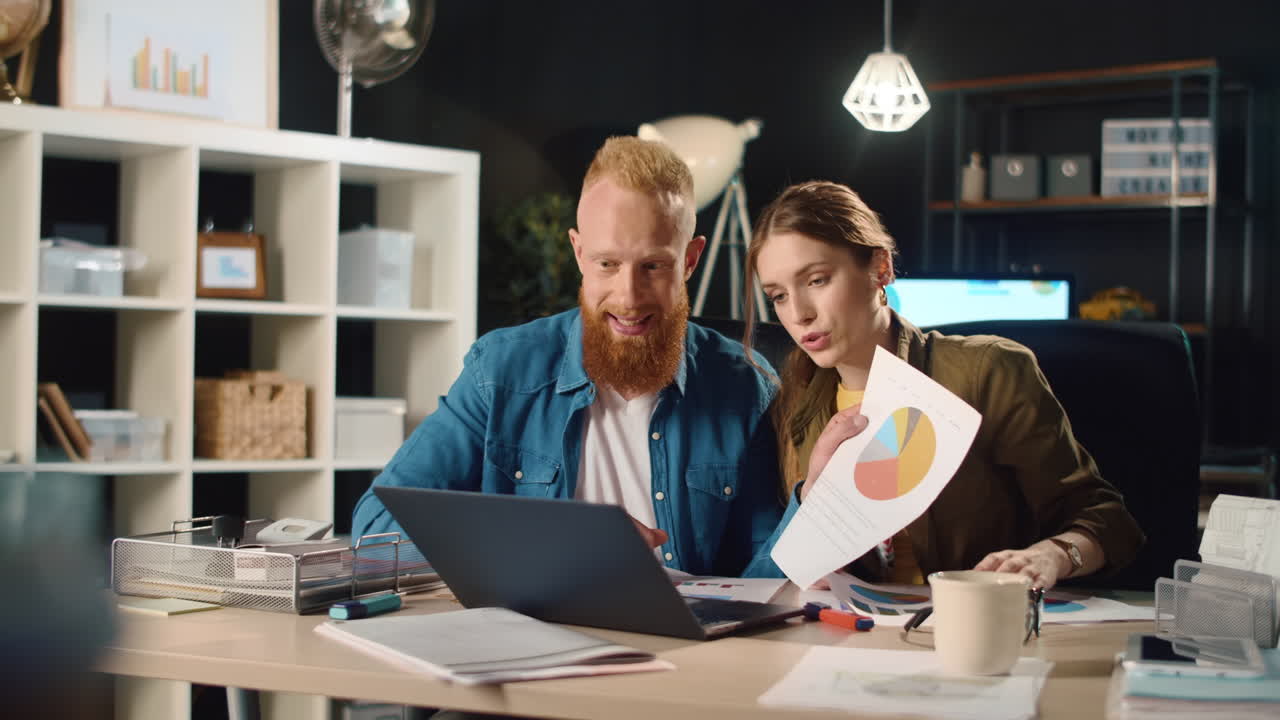 Friendly man and woman presenting project on video call in dark office.