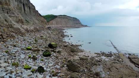 An-aerial-view-rises-up-from-a-rocky-beach-revealing-a-coastline-of-turquoise-sea-surrounding-high-cliffs-with-visible-strata-and-rockfall-that-are-topped-with-green-grass,-bushes-and-trees