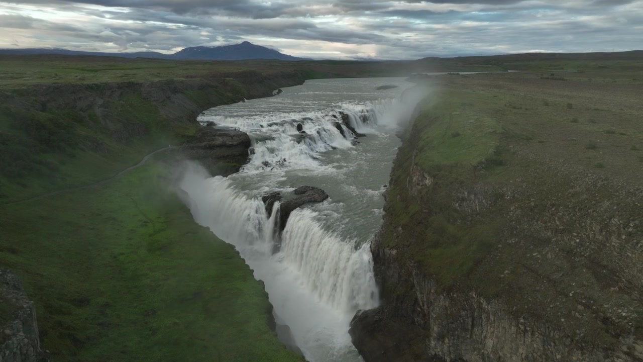 Une vue panoramique de la cascade de Gullfoss en Islande sous un ciel nuageux spectaculaire, entourée de plaines verdoyantes et de falaises escarpées
