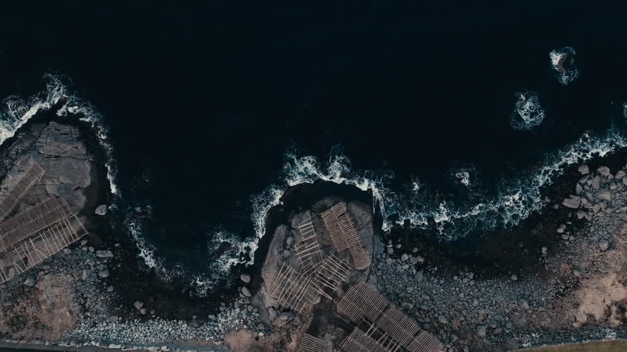 Lofoten Islands, Nordland, Norway - Rugged Arctic Coastline Meets Tradition, Where Ocean Waves Crash Beside Historic Fish Drying Racks - Aerial Ascend Shot