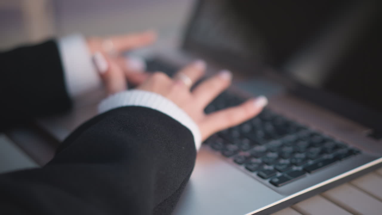 Close-up of hands adorned with polished nails and delicate rings typing on laptop keyboard, creating soft motion blur, cozy sweater sleeves enhance the scene with elegance