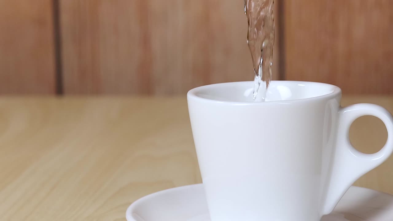 A steady stream of water fills a white ceramic cup placed on a saucer against a wooden backdrop.