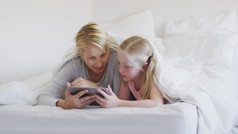 frontansicht einer kaukasischen frau und ihrer tochter, die ein digitales tablet auf dem bett benutzen