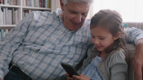 Abuelo-Feliz-Mostrando-A-La-Niña-Cómo-Usar-Un-Teléfono-Inteligente-Enseñando-A-Su-Nieta-Curiosa-Tecnología-Moderna-Niño-Inteligente-Aprendiendo-Teléfono-Móvil-Sentado-Con-El-Abuelo-En-El-Sofá