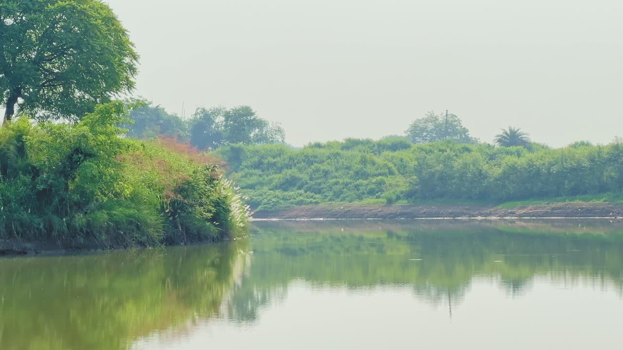 Still river water mirrors lush green banks and distant trees under a bright hazy sky, creating a soft serene landscape with gentle reflections along the bend