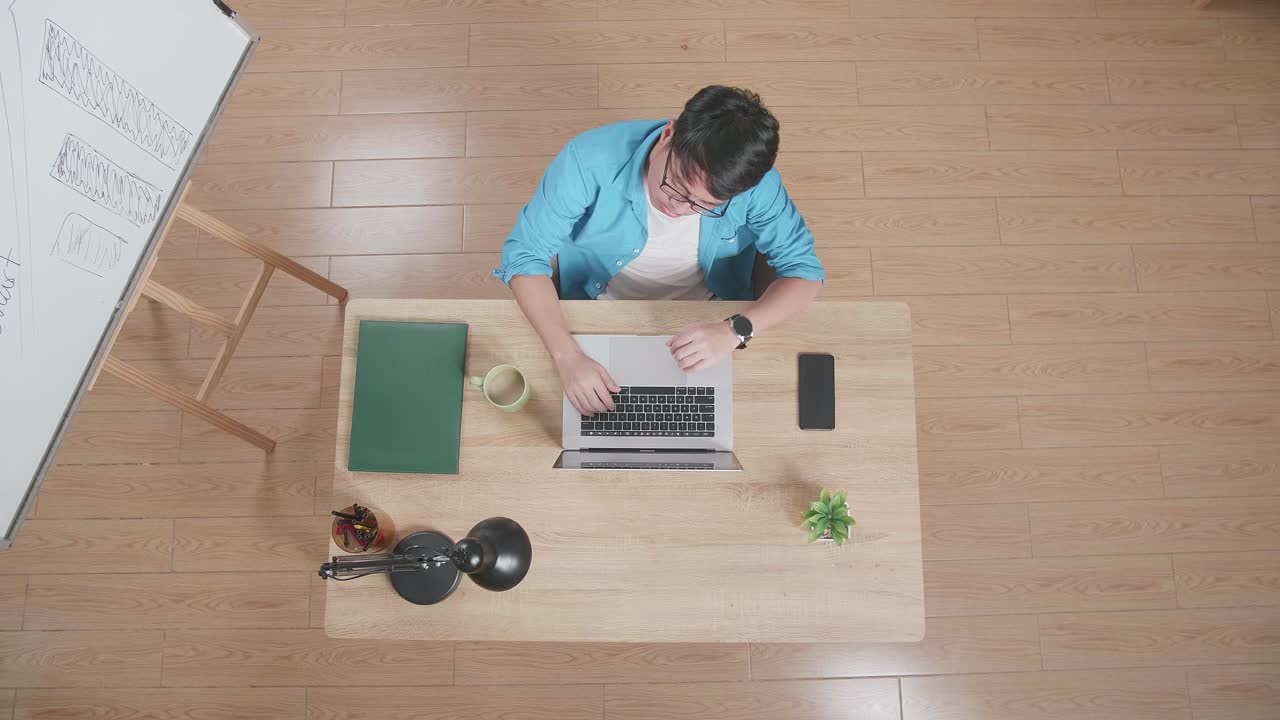 지친 남자가 머리가 아파서 스마트폰 에 노트북을 입력하고 집에 있는 화이트보드 에 나무 책상에 앉아 있습니다.