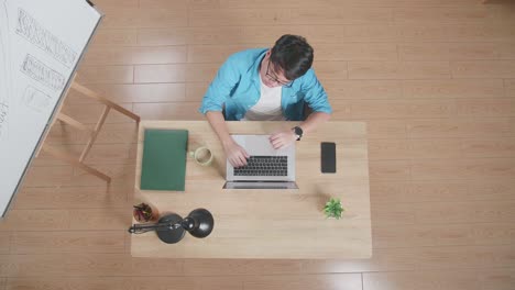 top view of a tired man having a headache typing on a laptop next to the smartphone while sitting at the wooden desk besides a whiteboard at home