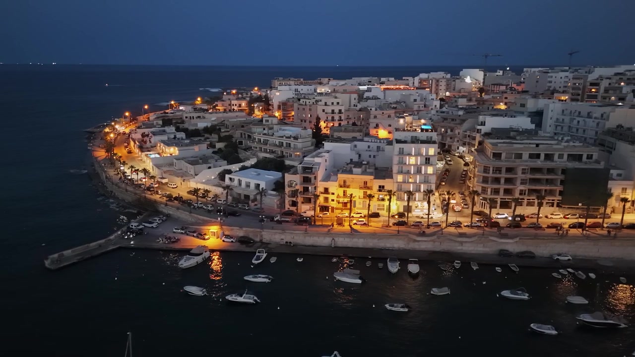 Lungomare di Marsaskala Bay, Malta, con barche, yacht e appartamenti illuminati lungo la passeggiata al tramonto. Porto mediterraneo e paesaggio urbano catturati da drone.