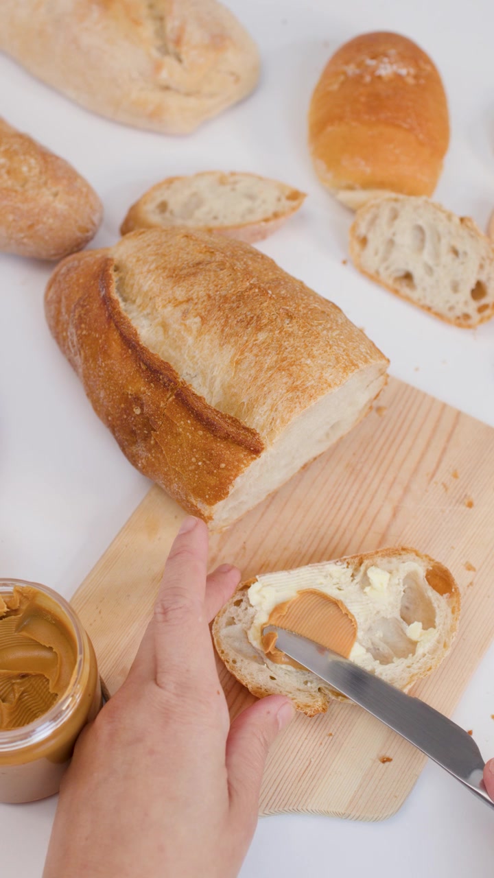Hands use a knife to spread peanut butter over buttered sourdough on a wooden board.