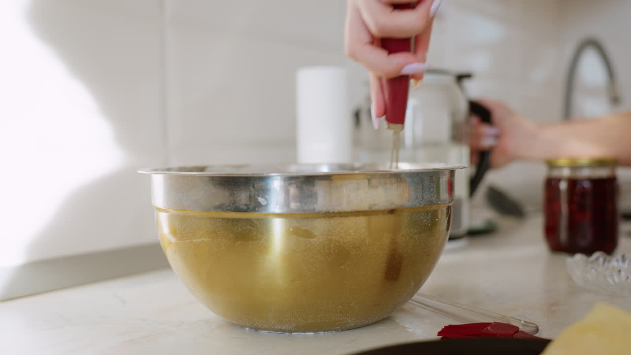 Close up of lady in green shirt pouring water from kettle into metal bowl while stirring pancake batter on kitchen counter with folded crepes on plate, sunlight casting shadow on white wall tiles